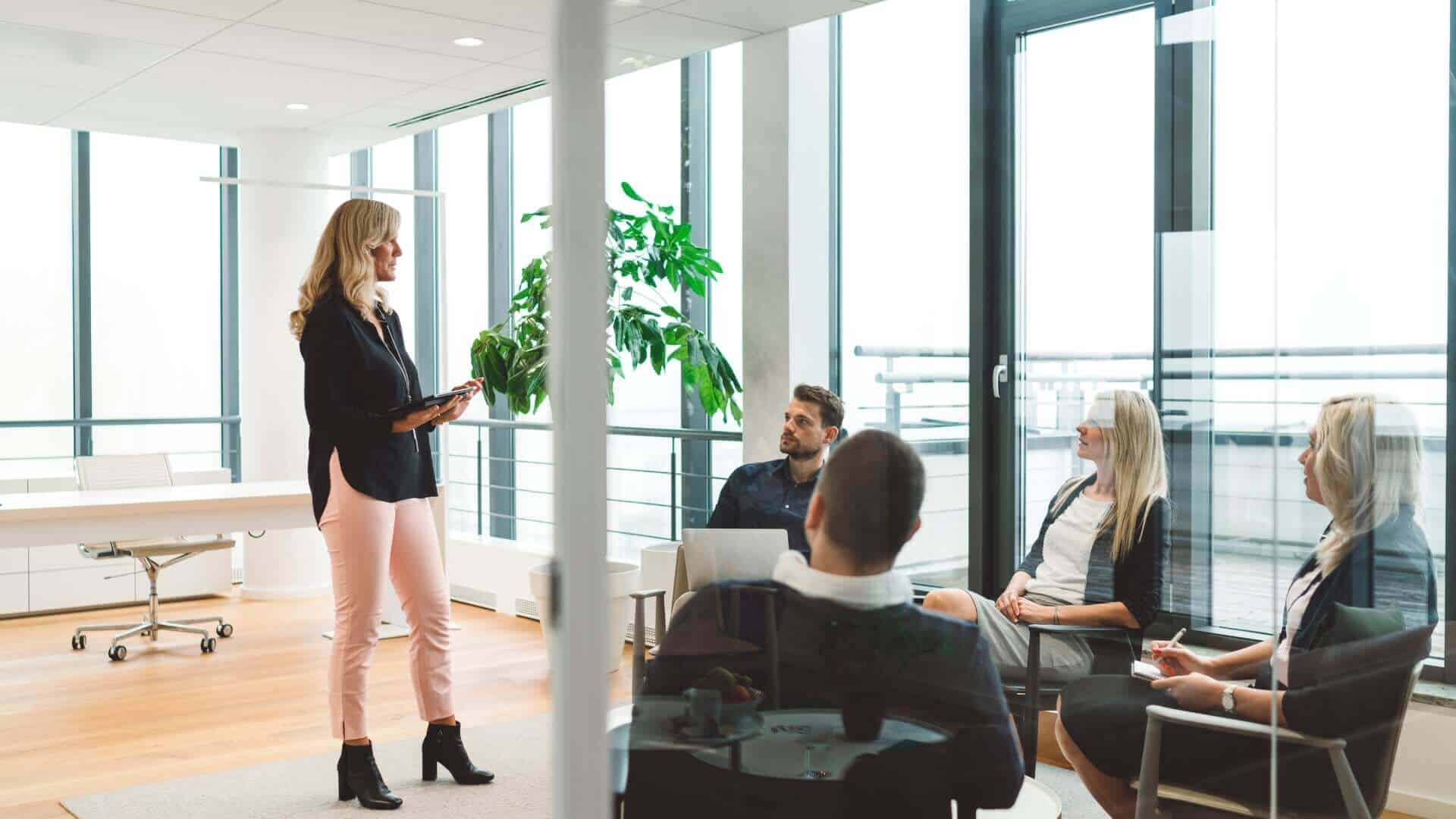 Eine Frau steht in einem modernen Büro und präsentiert vor einer Gruppe von Personen, die ihr zuhören.