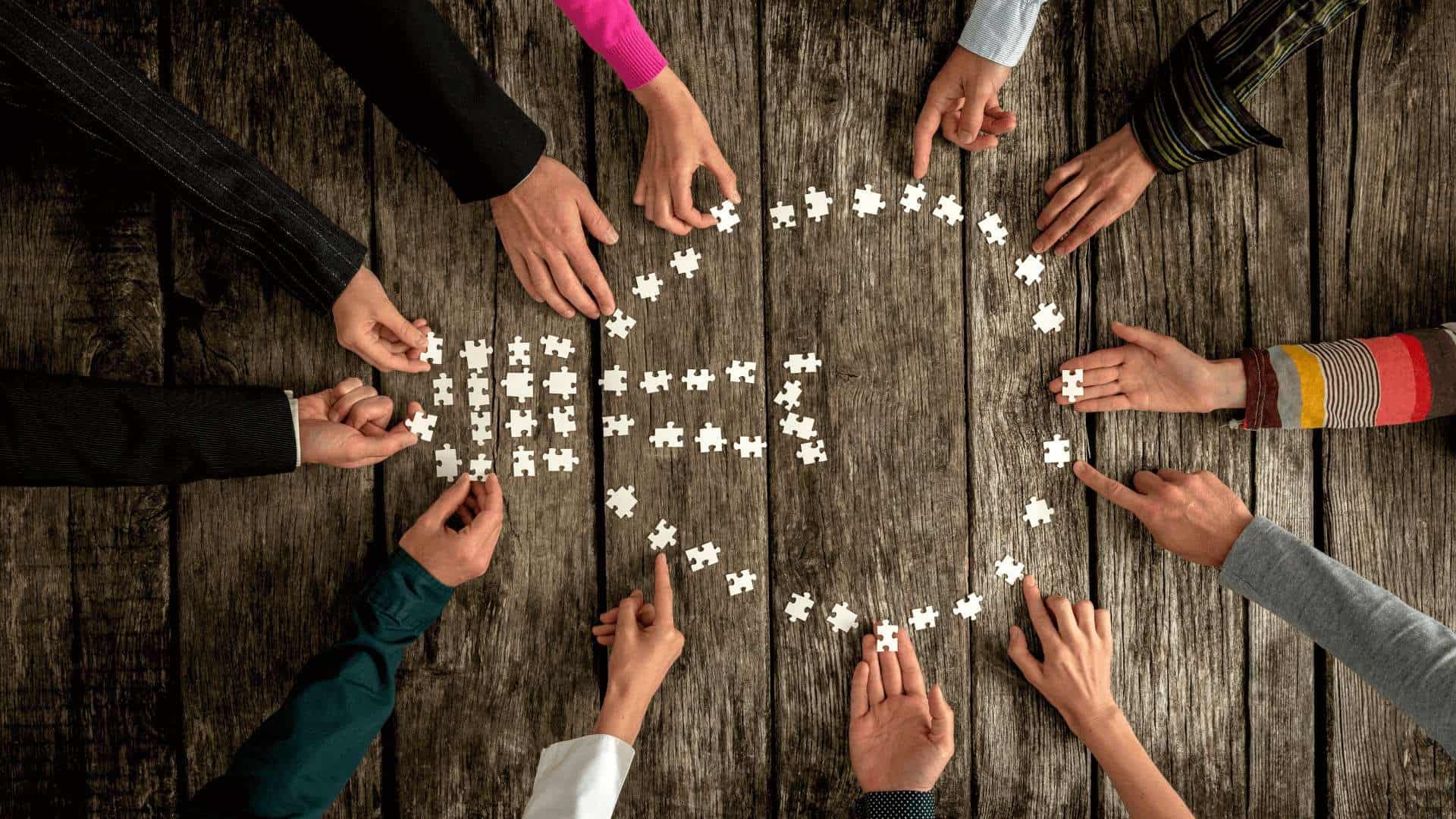 Mehrere Hände, die weiße Puzzlestücke auf einem Holztisch anordnen und zusammenfügen.