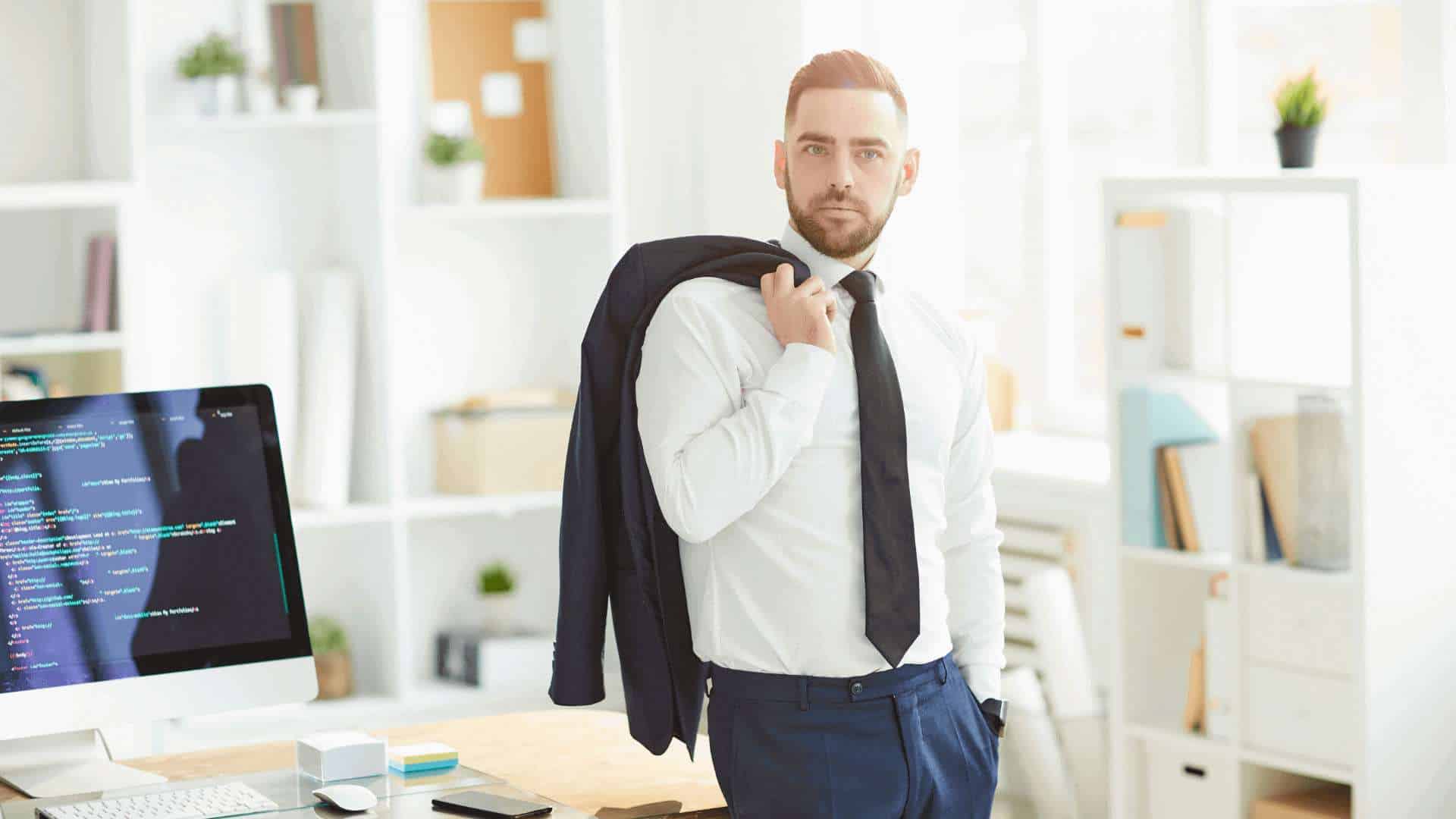 Mann in einem weißen Hemd und einer dunklen Krawatte hält sein Jackett in einem modernen Büro.